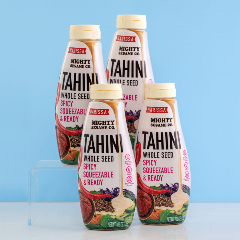 Four bottles of Mighty Sesame Harissa Tahini arranged against a light blue background, highlighting the squeezable design.