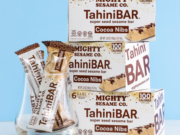 Boxes of Mighty Sesame Cocoa Nibs TahiniBARs stacked against a blue background, with individually wrapped bars in front.