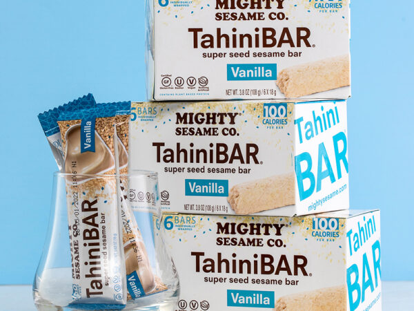 Boxes of Mighty Sesame Vanilla TahiniBARs stacked against a light blue background, with individually wrapped bars in front.