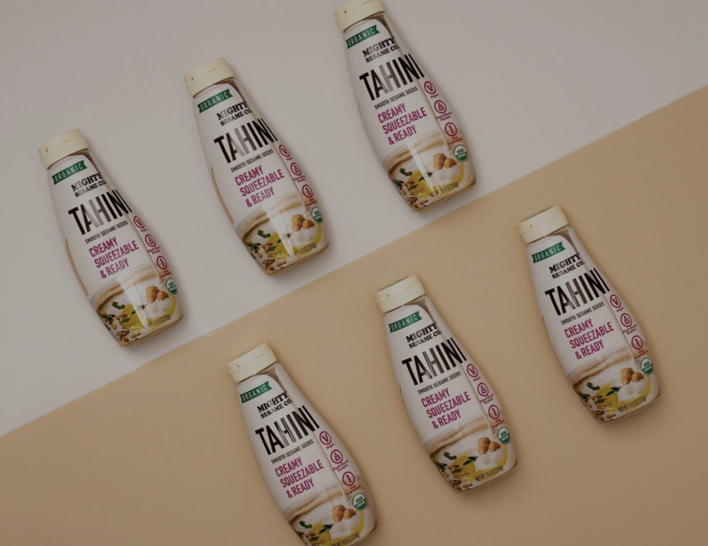 Six Mighty Sesame Organic Tahini squeeze bottles arranged in a grid pattern on a beige and white background.