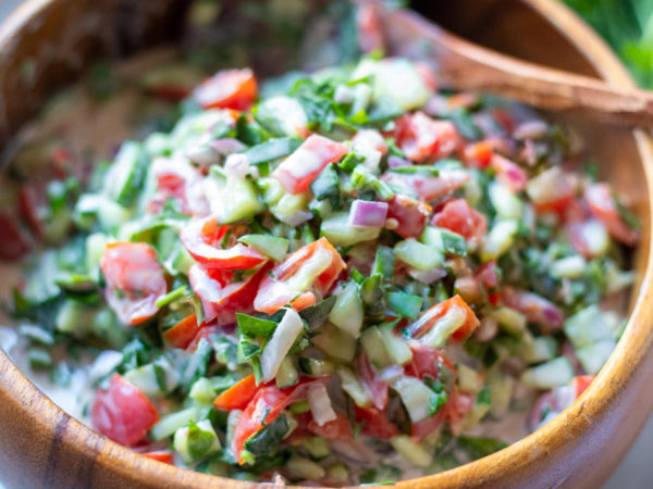 Chopped vegetable salad with tomatoes, cucumbers, and parsley, mixed with a creamy tahini dressing in a wooden bowl.