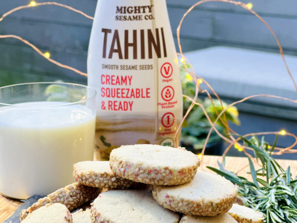 Golden tahini cookies arranged in a stack, with a bottle of Mighty Sesame Tahini and a glass of milk in the background.