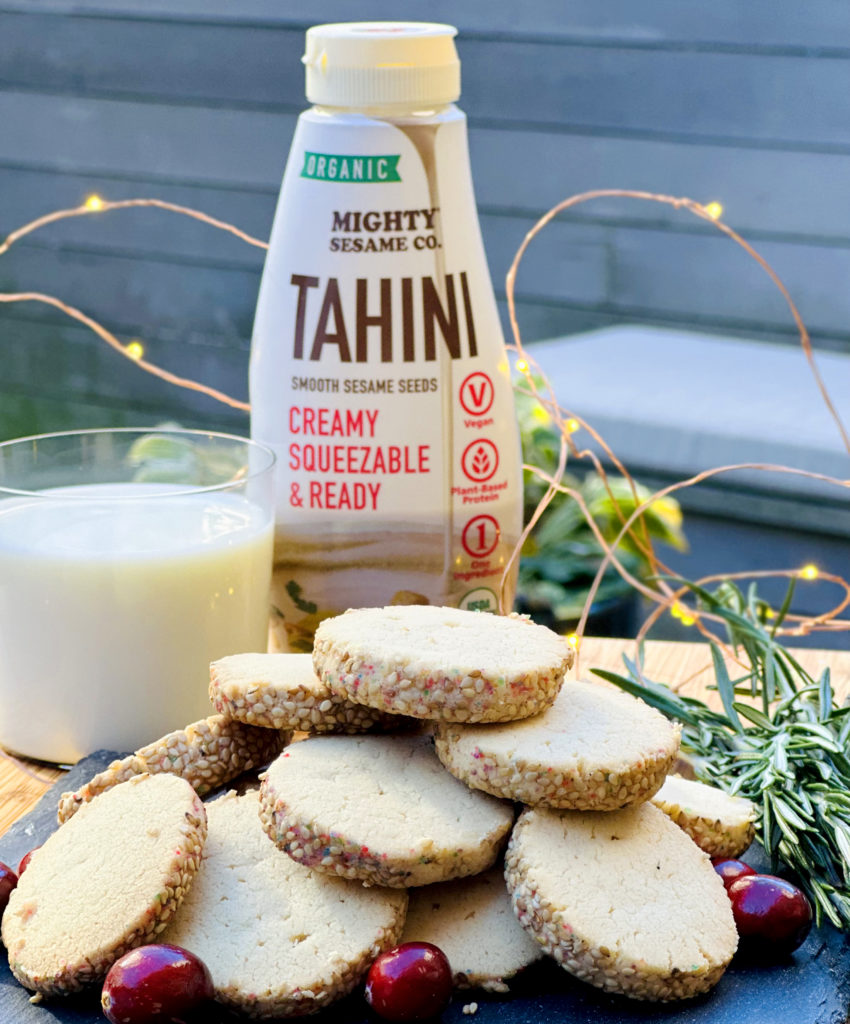 Golden tahini cookies arranged in a stack, with a bottle of Mighty Sesame Tahini and a glass of milk in the background.