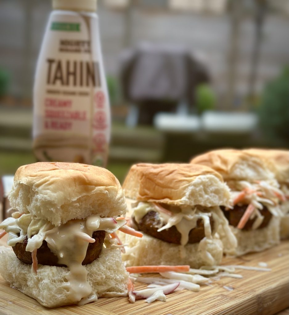 Three falafel sliders topped with fresh veggies and drizzled with tahini, served on mini buns.