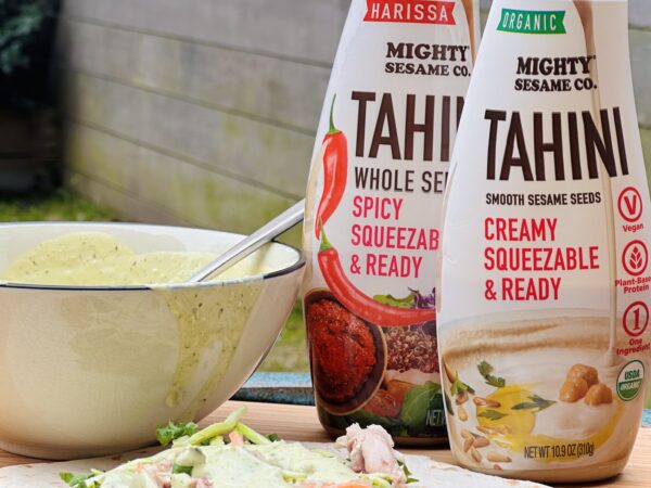 Two tahini squeeze bottles beside a bowl of green sauce and wrap fixings.