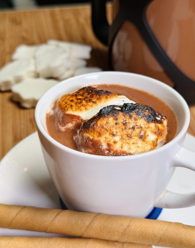 Cup of rich hot cocoa topped with toasted marshmallows and a drizzle of tahini, creating a nutty twist on a cozy beverage.