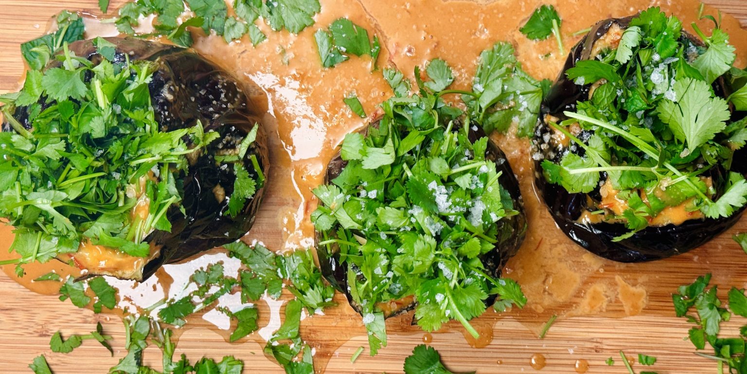 Roasted eggplant halves topped with fresh herbs like parsley and cilantro, drizzled with creamy tahini sauce.
