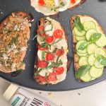 Three open-faced toasts with different hummus toppings, featuring a bottle of Mighty Sesame Tahini nearby.