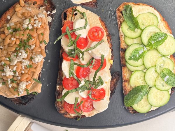 Three open-faced toasts with different hummus toppings, featuring a bottle of Mighty Sesame Tahini nearby.