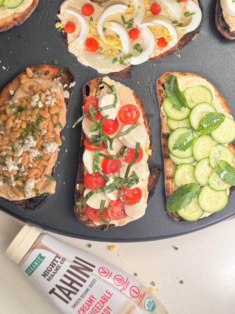 Three open-faced toasts with different hummus toppings, featuring a bottle of Mighty Sesame Tahini nearby.