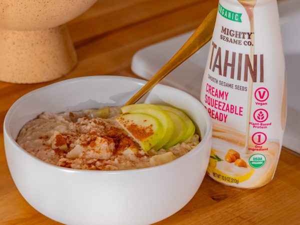 A bowl of tahini-based dish with nuts and green toppings, next to a bottle of Mighty Sesame Tahini on wood.