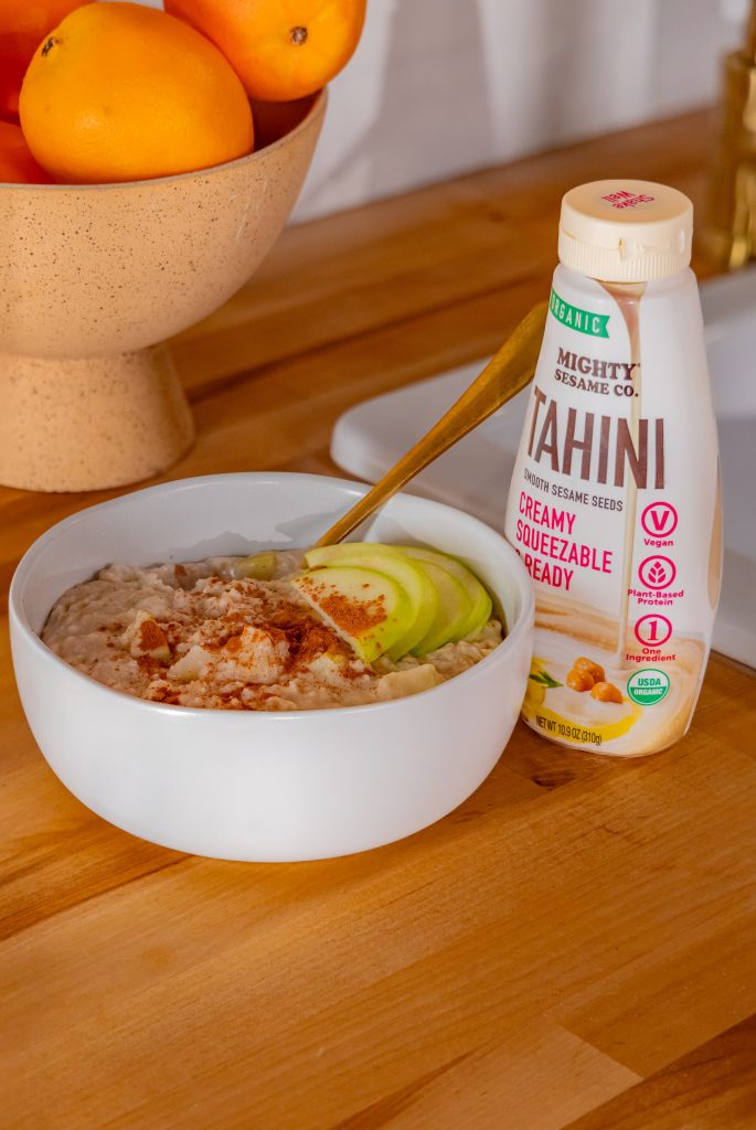 A bowl of tahini-based dish with nuts and green toppings, next to a bottle of Mighty Sesame Tahini on wood.