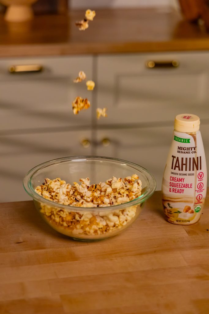 A bottle of Mighty Sesame tahini next to a bowl of assorted nuts with more falling in, showing preparation in progress.