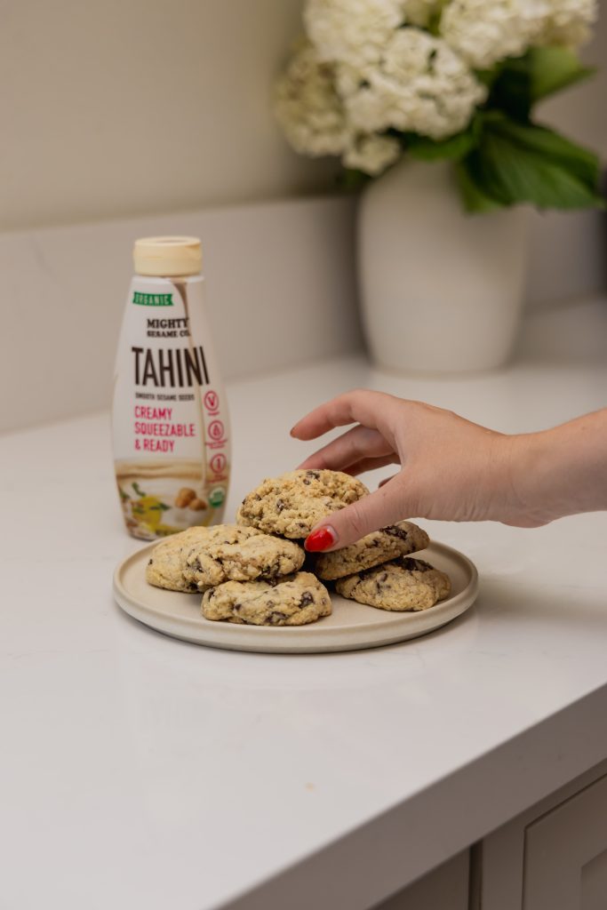 A bottle of Mighty Sesame Tahini cream is placed next to a plate of freshly baked cookies.