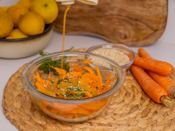 Mighty Sesame being poured into a bowl of grated carrots and mixed vegetables, with lemons and carrots in the background.