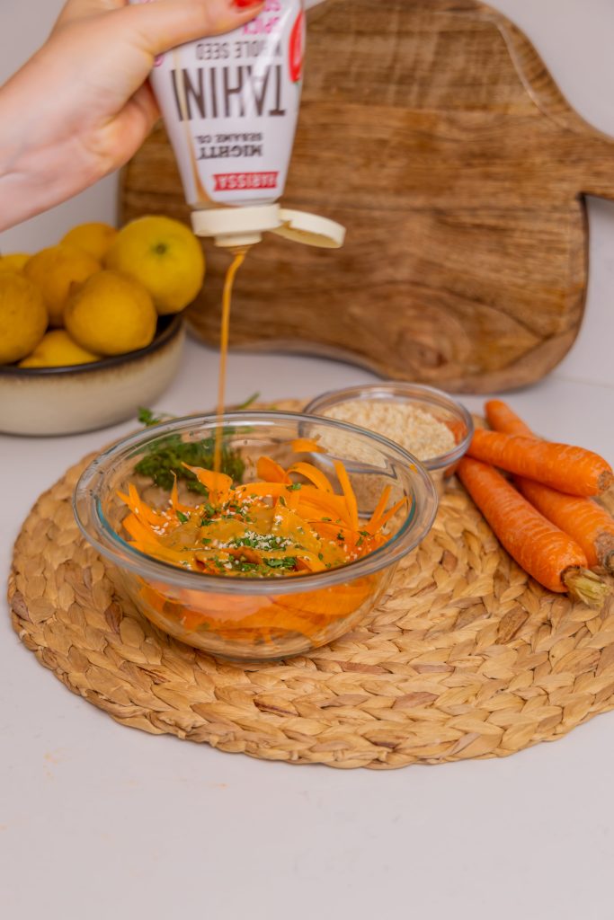 Mighty Sesame being poured into a bowl of grated carrots and mixed vegetables, with lemons and carrots in the background.