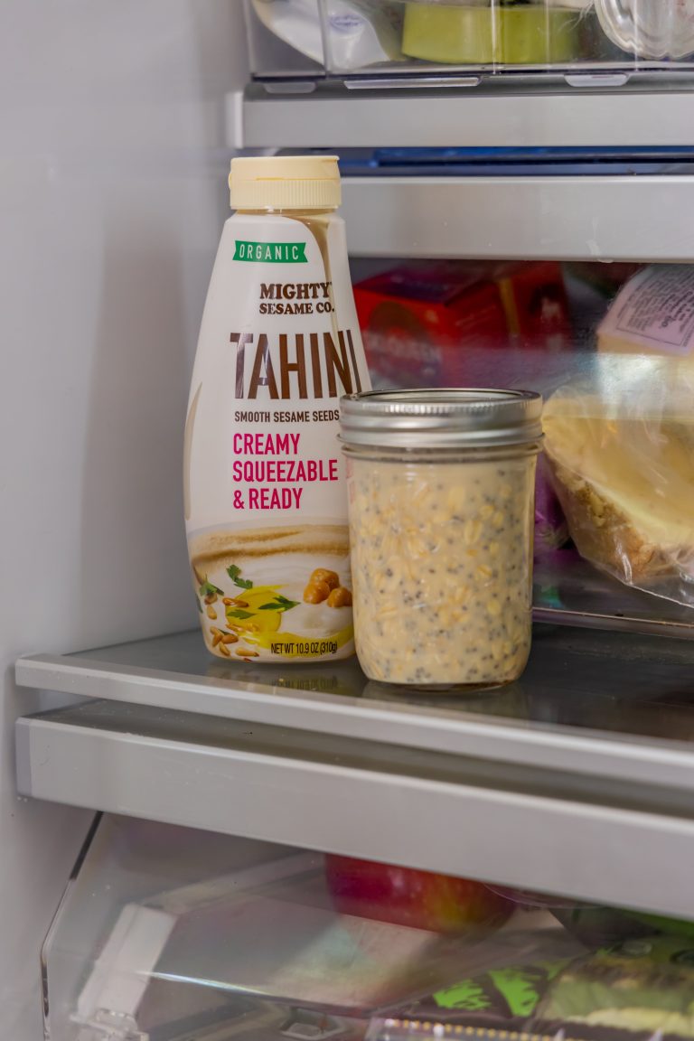 A bottle of Mighty Sesame tahini sits next to a jar of sesame seeds on a kitchen shelf with visible kitchenware.