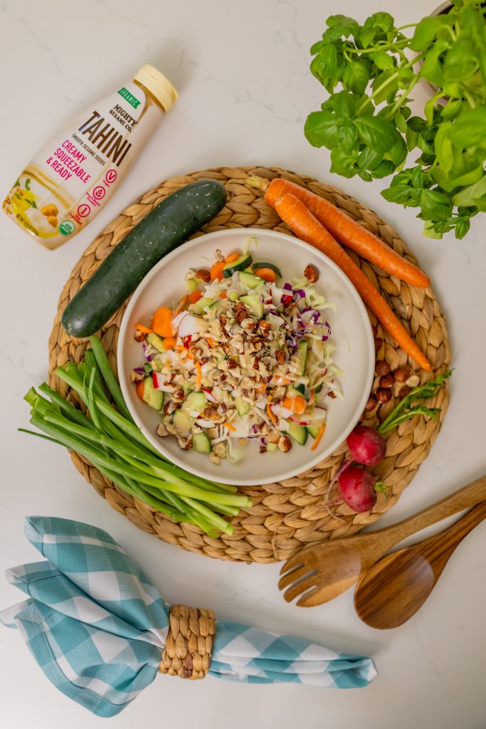 A vibrant bowl of salad with grains, herbs, and veggies, surrounded by fresh ingredients and Mighty Sesame tahini.