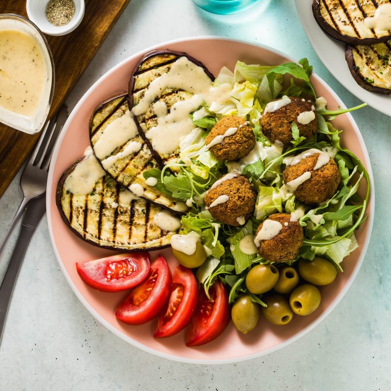 A fresh Mediterranean salad featuring grilled eggplant, falafel, olives, lettuce, tomato slices, and tahini dressing.