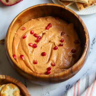 Wooden bowl of creamy sesame tahini, thick and smooth, topped with red seeds, set against a softly blurred background.