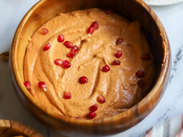 Wooden bowl of creamy sesame tahini, thick and smooth, topped with red seeds, set against a softly blurred background.