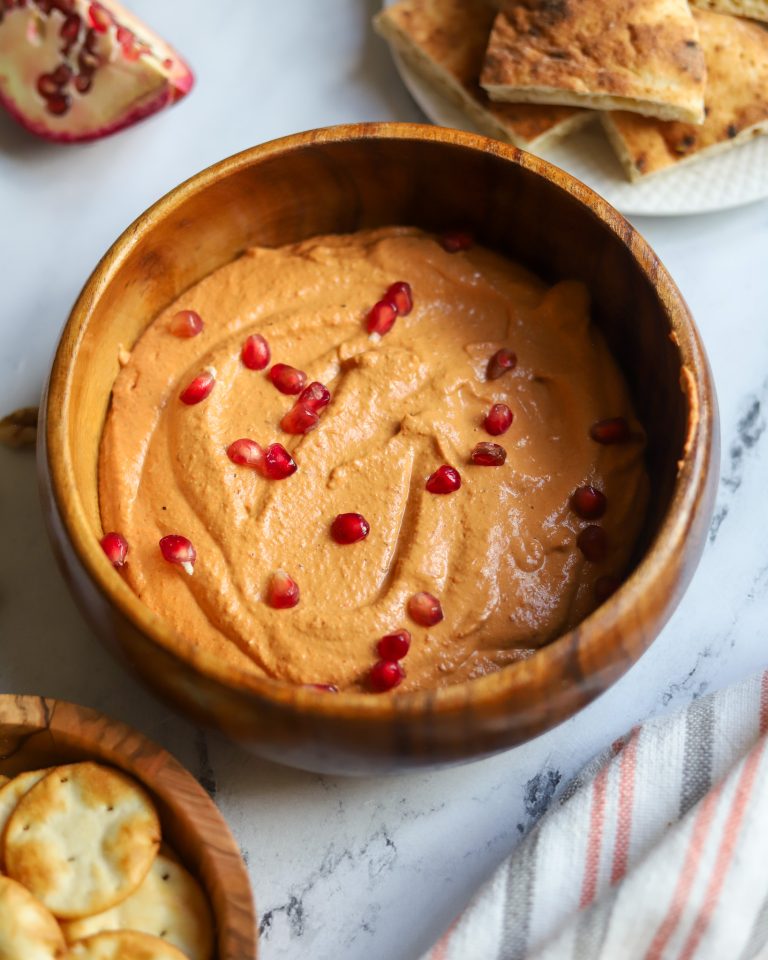Wooden bowl of creamy sesame tahini, thick and smooth, topped with red seeds, set against a softly blurred background.
