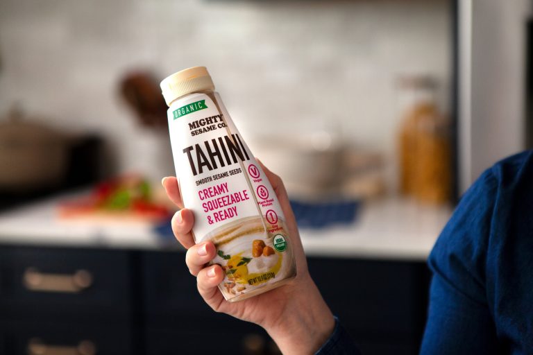 A person holds a bottle of Mighty Sesame Co. tahini, labeled "Creamy Squeezable & Ready," in a kitchen setting.