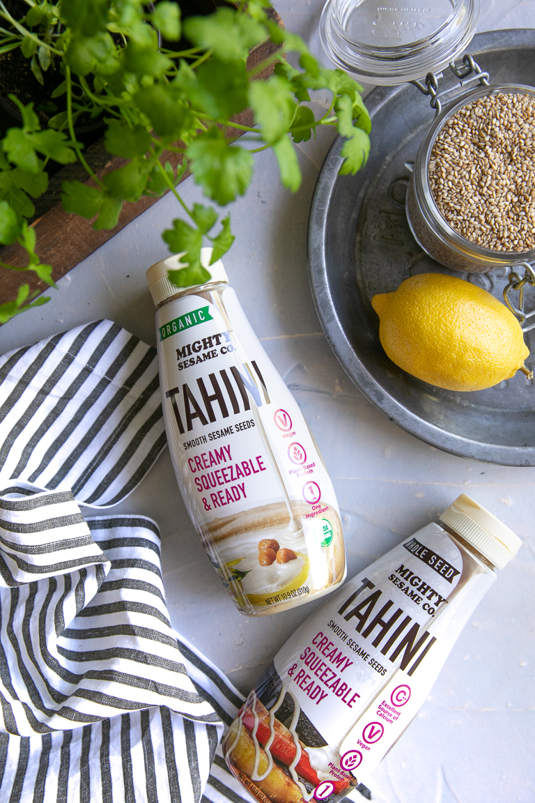 Two bottles of Mighty Sesame Co. tahini with a lemon, tahini seeds, and a potted herb on a textured surface.