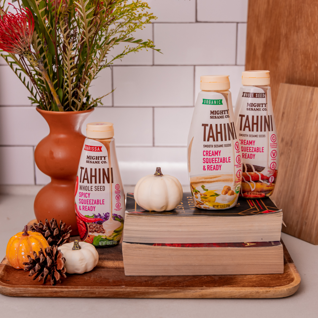 Mighty Sesame Co. tahini bottles (spicy and creamy) on books with decorative pumpkins and pinecones on a wooden tray.