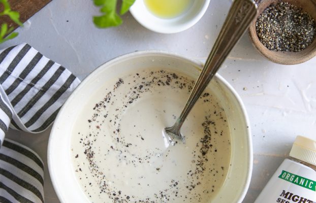 A bowl of creamy tahini topped with black pepper, surrounded by garlic, lemon, olive oil, spices, and fresh cilantro.