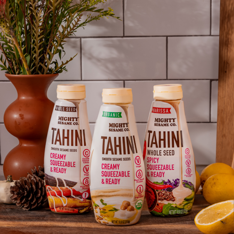 Three Mighty Sesame Co. Tahini squeeze bottles—Whole Seed, Organic, and Harissa Spicy—are displayed on a kitchen setup.