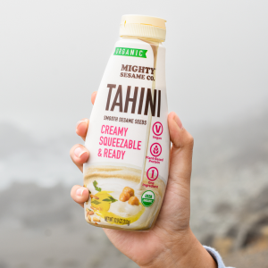 A hand holds a bottle of Mighty Sesame Co. Organic Tahini against a foggy outdoor background, showcasing its label.