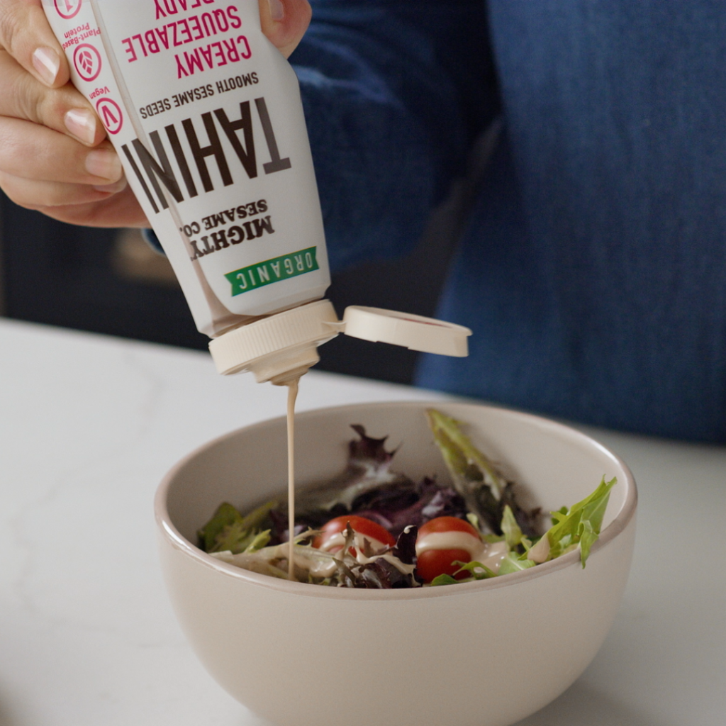 A person pours Mighty Sesame Co. Organic Tahini onto a fresh salad with cherry tomatoes in a beige bowl.