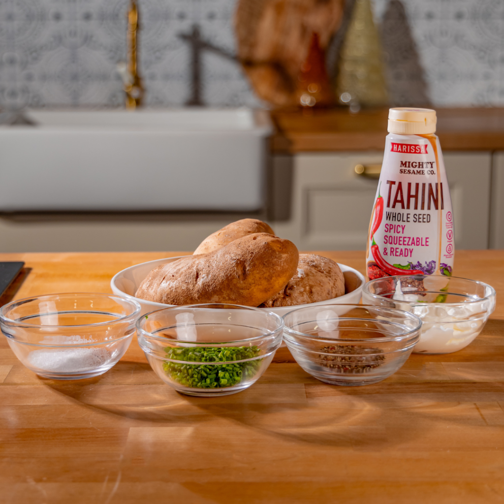 A kitchen counter with potatoes, seasoning bowls, and a bottle of spicy tahini, set against a stylish modern backdrop.