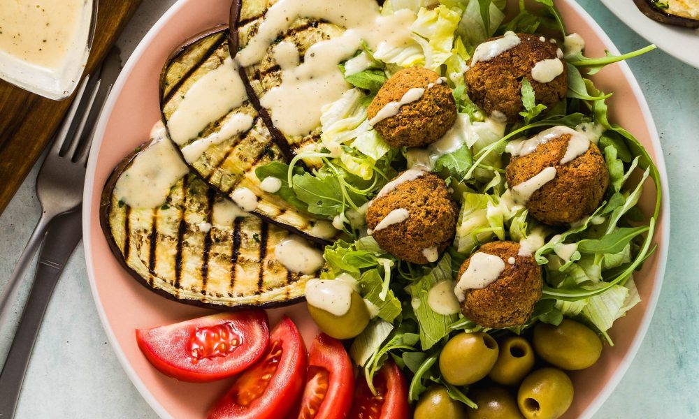 Falafel salad with grilled eggplant, tomatoes, and olives in a pink bowl with a white sauce.