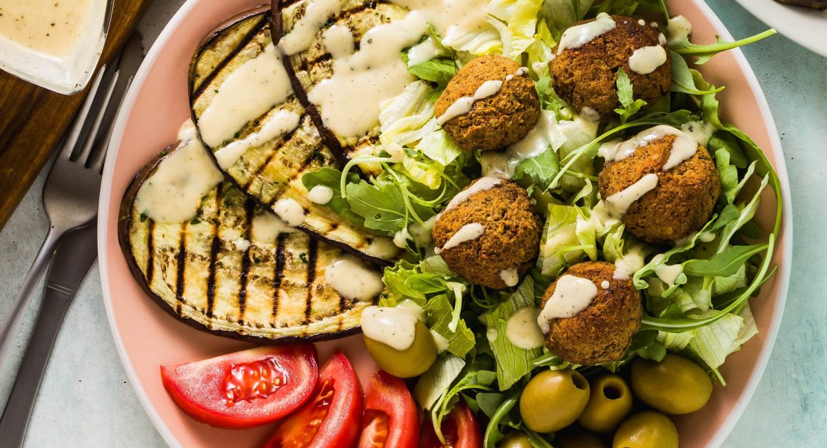 Falafel salad with grilled eggplant, tomatoes, and olives in a pink bowl with a white sauce.