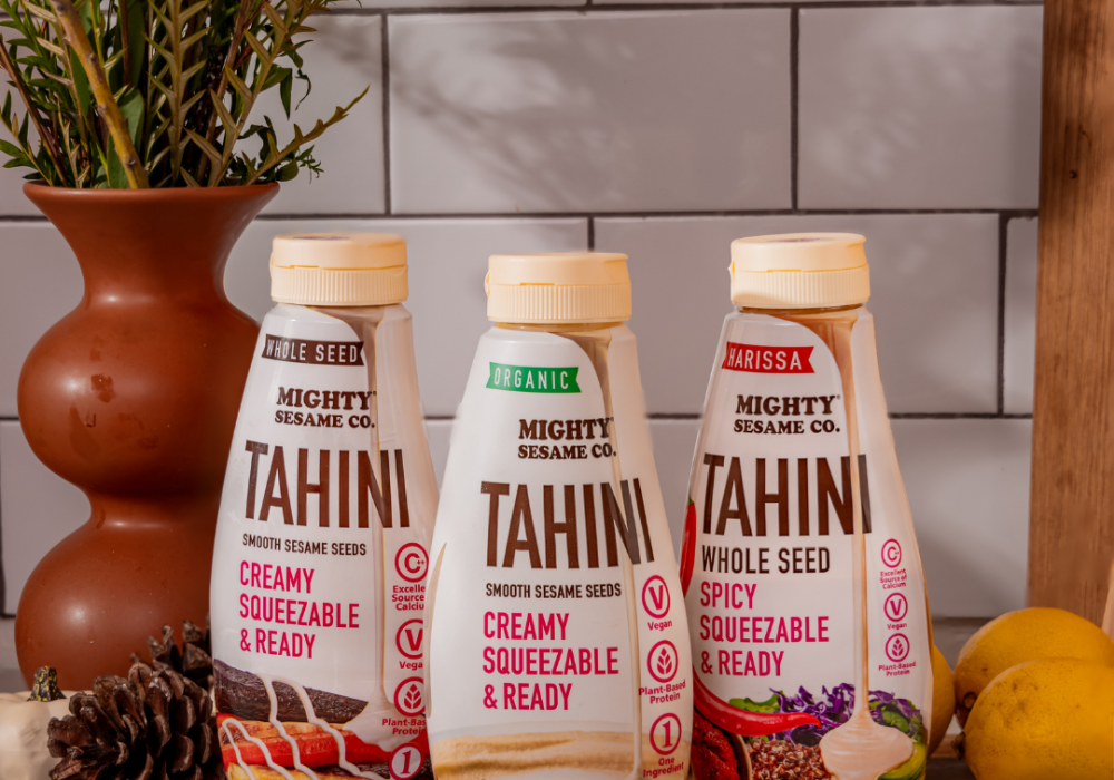 Three Mighty Sesame Co. Tahini squeeze bottles—Whole Seed, Organic, and Harissa Spicy—are displayed on a kitchen setup.