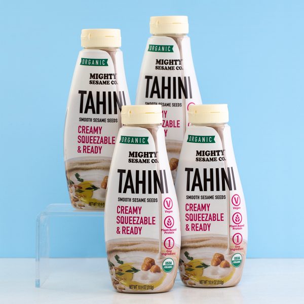 Four Mighty Sesame Co. organic tahini squeeze bottles arranged on a blue background.