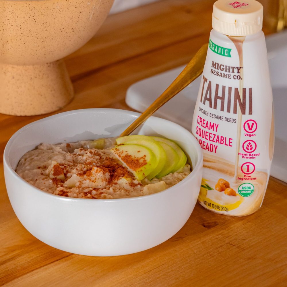 A bowl of tahini-based dish with nuts and green toppings, next to a bottle of Mighty Sesame Tahini on wood.