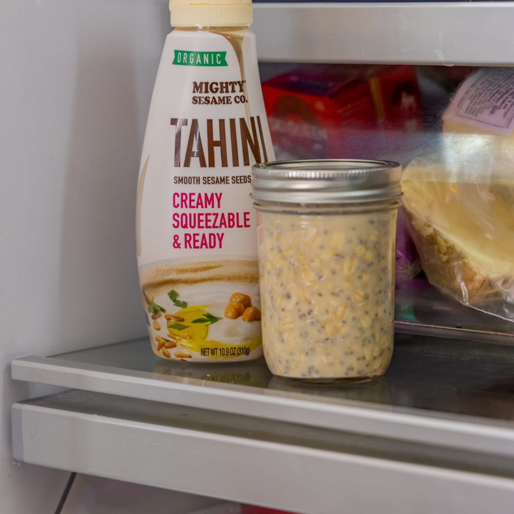 A bottle of Mighty Sesame tahini sits next to a jar of sesame seeds on a kitchen shelf with visible kitchenware.