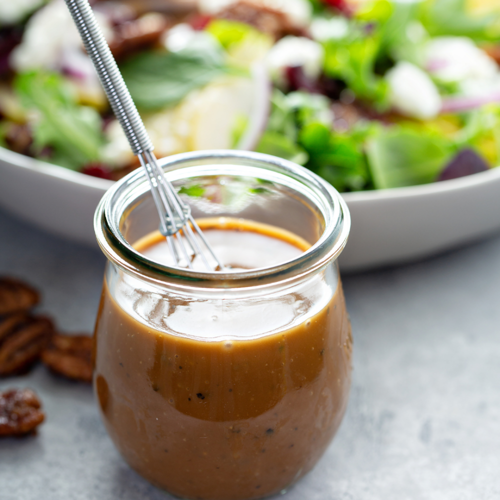 Glass jar of tahini balsamic dressing with a whisk, in front of a colorful salad in a white bowl with greens and onions.
