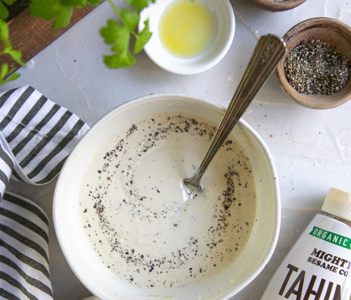 A bowl of creamy tahini topped with black pepper, surrounded by garlic, lemon, olive oil, spices, and fresh cilantro.